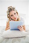 Pensive pretty blonde wearing hair curlers using tablet pc lying on cosy bed