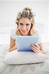 Cheerful pretty blonde wearing hair curlers using tablet pc lying on cosy bed