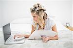 Cheerful cute blonde shopping online using laptop lying on cosy bed