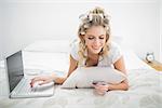 Smiling natural blonde shopping online using laptop lying on cosy bed