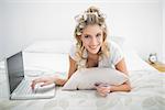 Cheerful natural blonde shopping online using laptop lying on cosy bed
