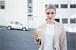 Serious stylish businesswoman holding coffee outside on urban background