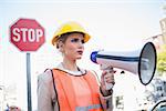 Frowning businesswoman wearing builders clothes holding megaphone outdoors on urban background