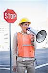 Angry businesswoman wearing builders clothes shouting in megaphone outdoors on urban background