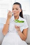 Relaxed pretty woman eating healthy salad sitting on sofa in bright living room