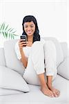 Happy black haired woman in white clothes typing on her mobile phone in a living room