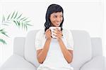 Pensive black haired woman in white clothes enjoying coffee in a living room