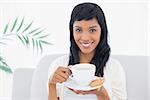 Calm black haired woman in white clothes drinking coffee in a living room