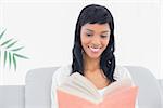 Thoughtful black haired woman in white clothes reading a book in a living room