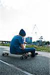 Backview of boy sitting on skateboard outdoors in an industrial area and looking at cell phone, Germany