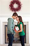 Happy young couple kissing in front of fireplace, holding Christmas present