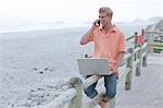 Man sitting on beach railing using phone and laptop