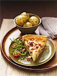 A slice of quiche lorraine with rocket salad and a side dish of potatoes