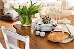 A bunch of white tulips in a glass vase next to a stack of plates, some wine glasses, decorative figs and a decorative artichoke