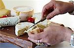 Slices of baguette being spread with Camembert, assorted types of cheese on a chopping board