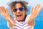 A beautiful and happy mixed race African American female girl child wearing sunglasses in bright sunshine on vacation in front of a blue tropical sea laughing & reaching to camera