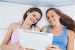 Friends looking at tablet and laughing lying on bed at home at sleepover