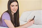 Cheerful brunette looking at camera and lying on a bed using a mobile phone in a bedroom