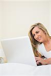 Happy mature blonde woman typing on a laptop lying on her bed in a bedroom