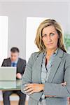 Serious blonde businesswoman looking at camera crossed arms with a mature businessman working on laptop on background at office