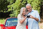 Cheerful mature couple looking at pictures on their camera while leaning against their cabriolet