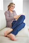 Smiling blonde woman lying on a sofa in a living room concentrated reading a book