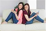 Two smiling teenage girls sitting back to back on a sofa using a tablet pc and a mobile phone