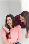 Smiling brunette giving a present to her friend who is using a mobile phone sitting on a sofa in a living room