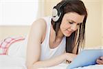 Smiling brunette listening to music with a headset and using a tablet pc lying on a bed in a bedroom