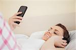 Happy brunette listening to music with a headset and using a mobile phone lying on a bed in a bedroom