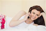 Smiling brunette calling someone with a mobile phone lying on a bed in a bedroom