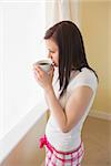 Thoughtful brunette drinking a cup of coffee standing in a bedroom