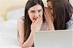 Happy girl telling a secret to her friend in front of laptop at home in bedroom