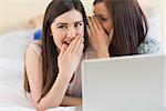 Smiling girl telling a secret to her friend in front of laptop at home in bedroom