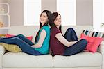 Two smiling friends sitting back to back on the sofa at home in living room