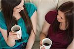 Two smiling friends having coffee and chatting on the couch at home in the living room