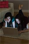 Two friends lying on floor using laptop together in the dark at home in the living room