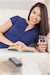 Pretty asian girl lying on the sofa reading a novel and drinking water at home in the sitting room