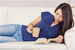 Beautiful asian girl lying on the sofa reading a novel and drinking hot beverage in living room at home