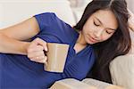 Attractive asian girl lying on the sofa reading a novel and drinking hot beverage in living room at home