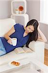 Pretty young asian woman lying on the sofa reading a book holding her coffee in living room at home