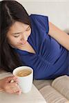 Young asian woman lying on the sofa reading a book holding her coffee in living room at home