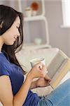 Young asian woman sitting on the sofa reading a book holding her coffee in living room at home