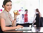 Happy businesswoman holding wine glass using laptop and looking at camera in a restaurant