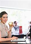 Smiling businesswoman using laptop while holding wine glass looking at camera