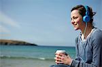 Young woman at coast with coffee and earphones