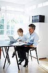 Man sitting at desk with son on his knee with laptop computer and paperwork