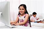 Girl using a computer with her brother reading a book in the background