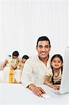 South Indian man using a desktop computer with his daughter with other family in the background