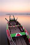 Traditional rowing boat moored on the edge of flat calm Taungthaman Lake at dawn with the colours of the sky reflecting in the calm water, close to the famous U Bein teak bridge, near Mandalay, Myanmar (Burma), Asia